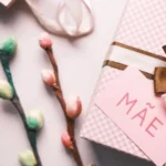 Mão escrevendo um cartão de aniversário personalizado para a mãe, com flores e uma xícara de café ao fundo, simbolizando carinho e dedicação.