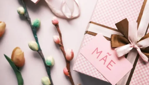 Mão escrevendo um cartão de aniversário personalizado para a mãe, com flores e uma xícara de café ao fundo, simbolizando carinho e dedicação.
