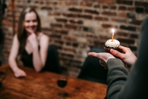 Casal sorrindo e abraçando em festa de aniversário, com balões e bolo em segundo plano, representando amor e celebração