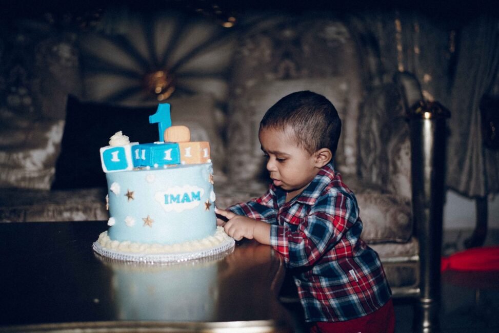 Pais felizes segurando as mãos de um bebê de 1 ano soprando a primeira vela em um bolo de aniversário, com balões ao fundo.