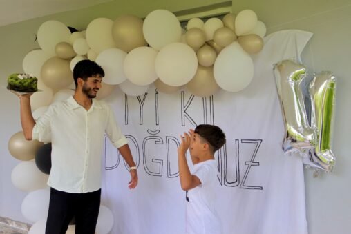 Mãe e filho sorrindo e abraçando em uma festa de aniversário, com balões e bolo ao fundo, capturando um momento de carinho.