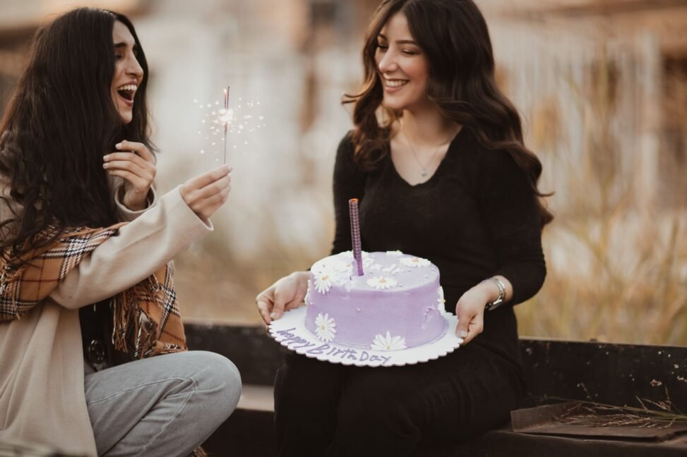 Duas irmãs sorrindo e se abraçando em uma foto de aniversário, com balões ao fundo.
